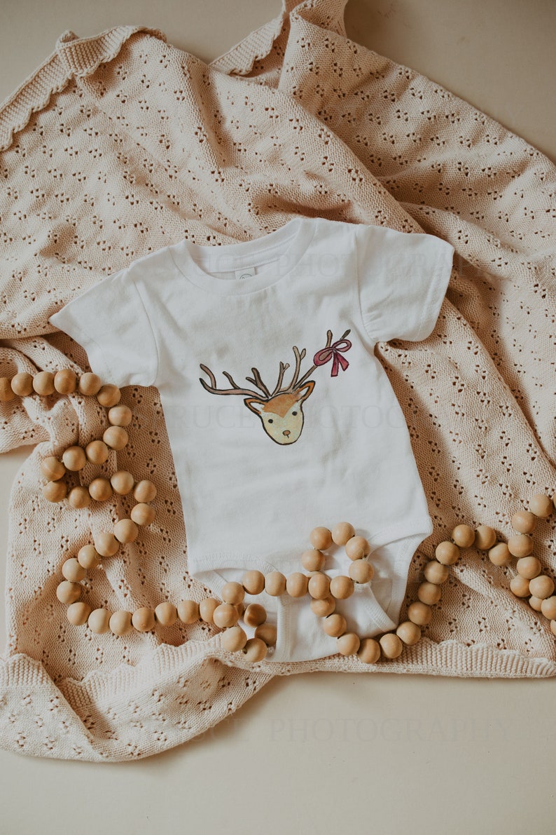 May include: A white baby onesie with a brown and red reindeer illustration. The onesie is lying on a beige knitted blanket with wooden beads.