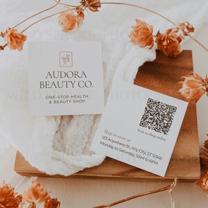Może przedstawiać: Biała wizyt&oacute;wka z tekstem "Audora Beauty Co." i "One-Stop Health & Beauty Shop". Wizyt&oacute;wka zawiera r&oacute;wnież kod QR z tekstem "Scan to shop online" i adresem "123 Anywhere St, Any City, ST 12345". Wizyt&oacute;wka leży na drewnianej powierzchni z suszonymi kwiatami.