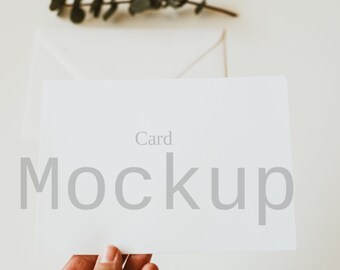 May include: A white card mockup with the word "Card" above the word "Mockup" in gray. A hand holds the card, with a white envelope and sprig of greenery in the background. The image has a soft, neutral color palette.