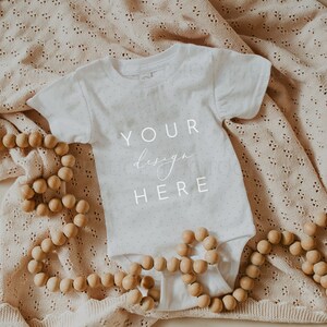 May include: A white baby onesie with the text "YOUR design HERE" on a beige knitted blanket with wooden beads.
