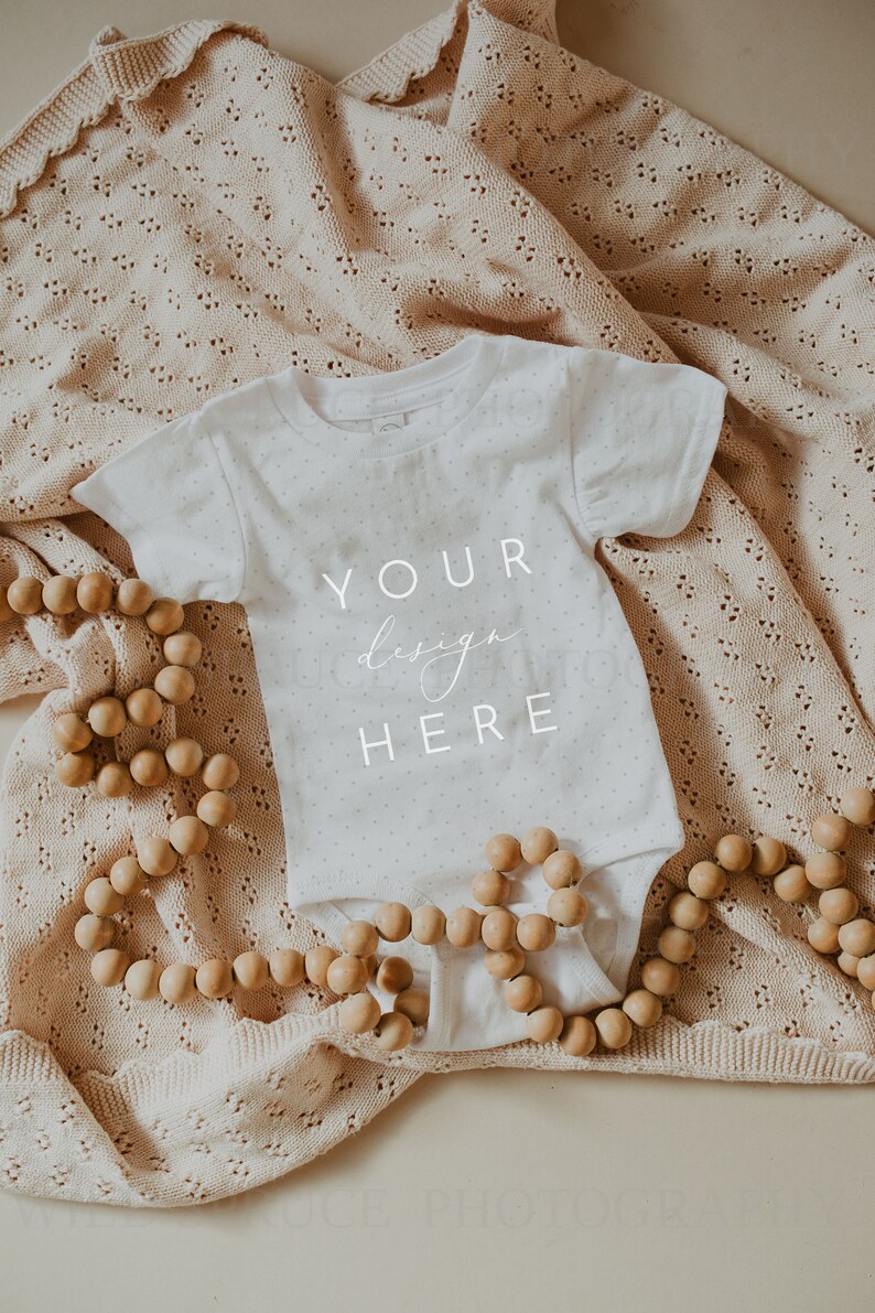 May include: A white baby onesie with the text "YOUR design HERE" on a beige knitted blanket with wooden beads.