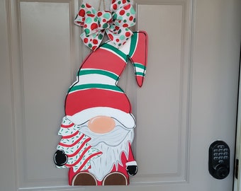 May include: A wooden Christmas gnome door hanger. The gnome is red, white, and green with a long hat and a white beard. It holds a Christmas tree and is topped with a polka dot bow.
