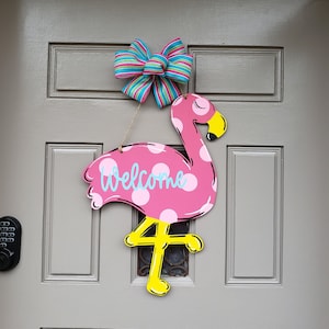 May include: Pink flamingo door hanger with white polka dots and a yellow beak. The flamingo has the word "Welcome" written on its back and a yellow number "4" on its leg. A blue, green, and pink bow is hanging above the flamingo.