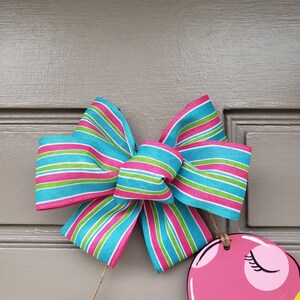 May include: A pink flamingo door hanger with a blue, green, and pink striped bow. The bow is tied with twine.