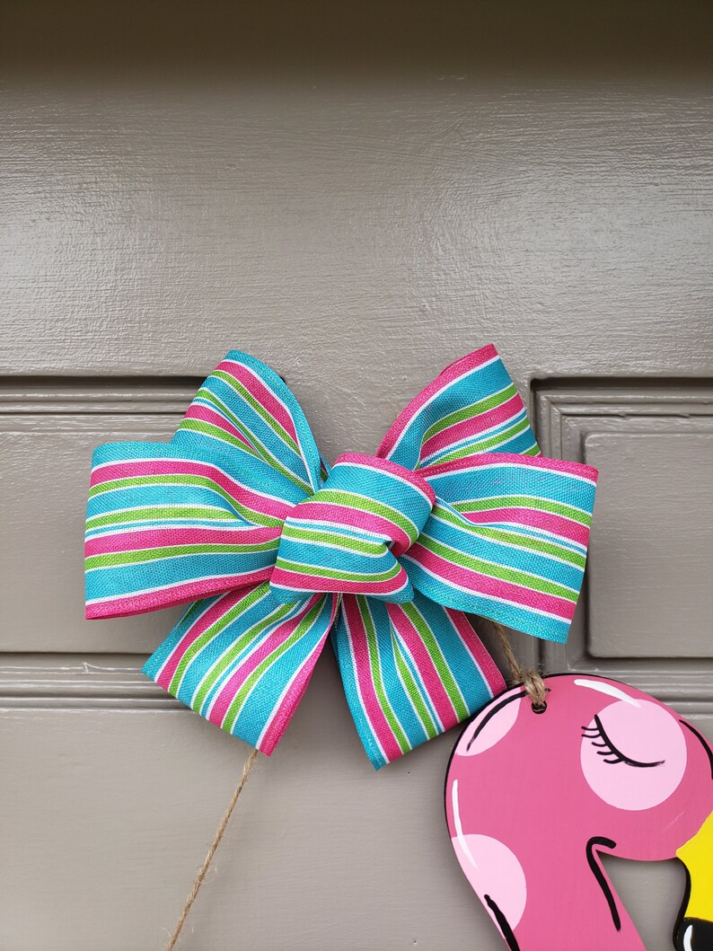 May include: A pink flamingo door hanger with a blue, green, and pink striped bow. The bow is tied with twine.