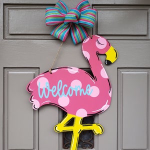 May include: Pink flamingo door hanger with white polka dots and the word "Welcome" in blue. The flamingo has yellow legs and a yellow beak. A blue and green striped bow is attached to the top of the hanger.