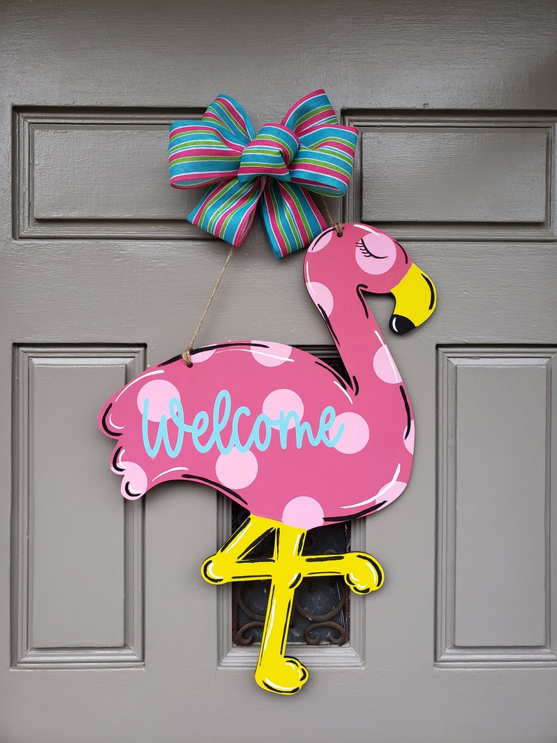 May include: Pink flamingo door hanger with white polka dots and the word "Welcome" in blue. The flamingo has yellow legs and a yellow beak. A blue and green striped bow is attached to the top of the hanger.