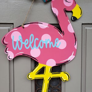 May include: Pink flamingo door hanger with white polka dots and the word "Welcome" in blue. The flamingo has yellow legs and a yellow beak.