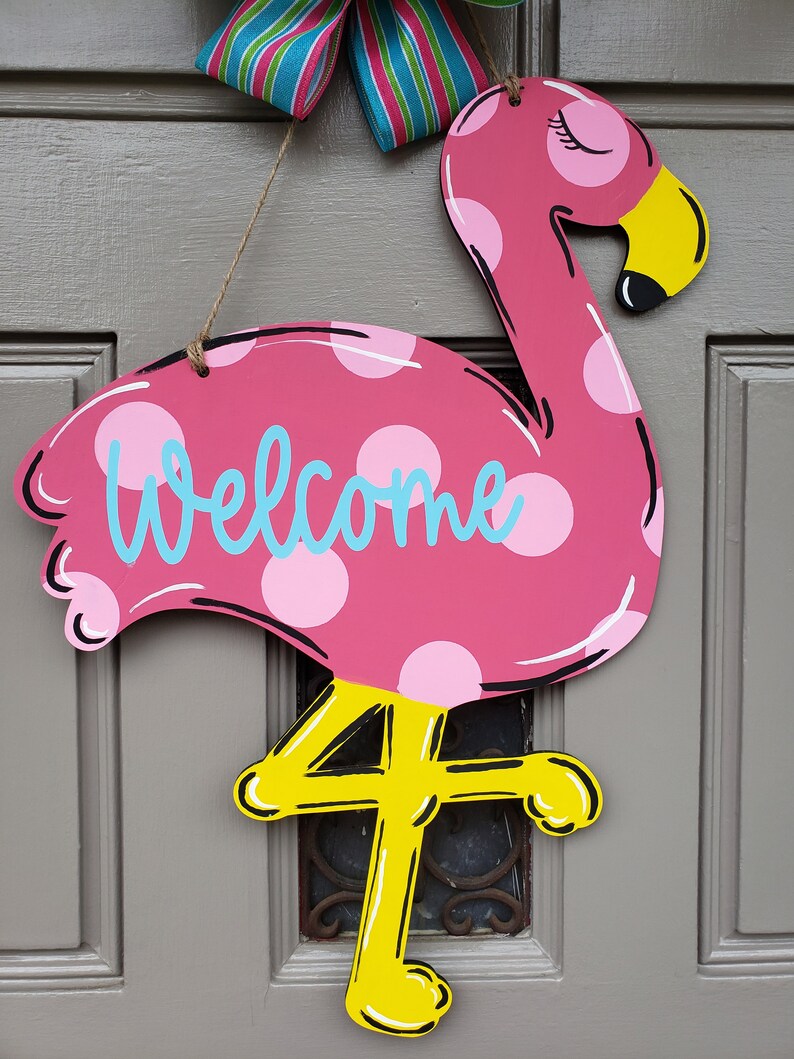 May include: Pink flamingo door hanger with white polka dots and the word "Welcome" in blue. The flamingo has yellow legs and a yellow beak.