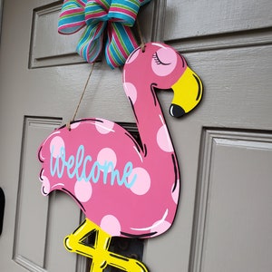 May include: Pink flamingo door hanger with white polka dots and the word "welcome" in blue. The flamingo has yellow legs and a yellow beak. A blue and pink striped bow is hanging above the flamingo.
