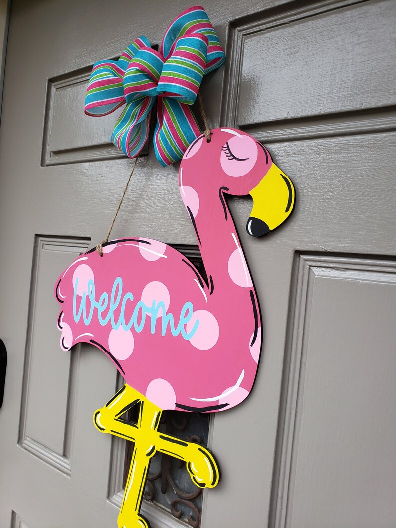 May include: Pink flamingo door hanger with white polka dots and the word "welcome" in blue. The flamingo has yellow legs and a yellow beak. A blue and pink striped bow is hanging above the flamingo.