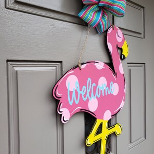 May include: Pink flamingo door hanger with white polka dots and the word "Welcome" written in blue. The flamingo has a yellow beak and legs, and the number "4" is painted on the leg.