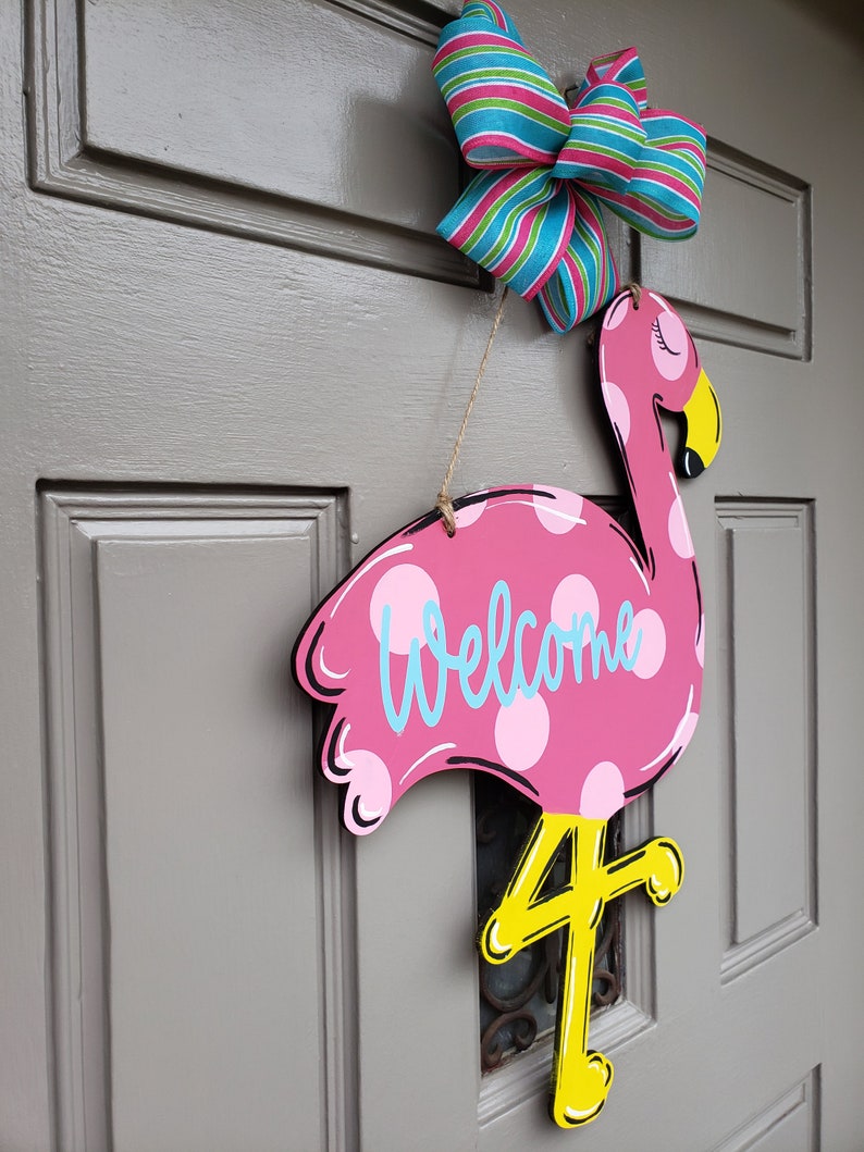 May include: Pink flamingo door hanger with white polka dots and the word "Welcome" written in blue. The flamingo has a yellow beak and legs, and the number "4" is painted on the leg.