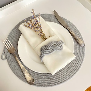 May include: A place setting with a white plate, a folded white napkin held by a gray rope knot napkin ring, and a sprig of lavender. A gray placemat, fork, and knife complete the elegant table setting.