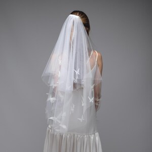 May include: A white bridal veil with butterfly accents. The veil is made of a sheer fabric and features delicate butterfly appliqu&eacute;s. The bride is wearing a white dress. The veil is secured with a hair accessory.