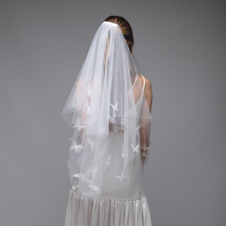 May include: A white bridal veil with butterfly accents. The veil is made of a sheer fabric and features delicate butterfly appliqu&eacute;s. The bride is wearing a white dress. The veil is secured with a hair accessory.