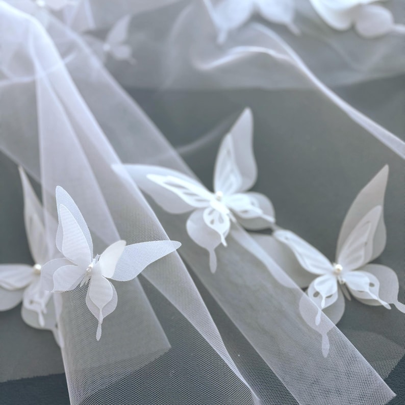 May include: Close-up of white butterfly appliqu&eacute;s on a sheer fabric. The butterflies have delicate, layered wings and are adorned with small pearl accents. The fabric is draped, creating soft folds and shadows.