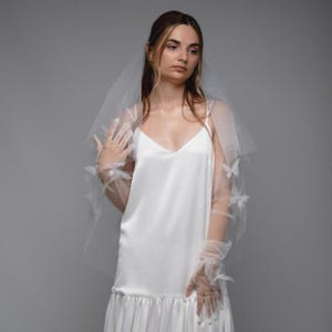 May include: A white wedding dress with a V-neckline and a tiered skirt. The bride is wearing a sheer veil adorned with butterfly accents and matching gloves. The dress is made of a smooth, flowing fabric.