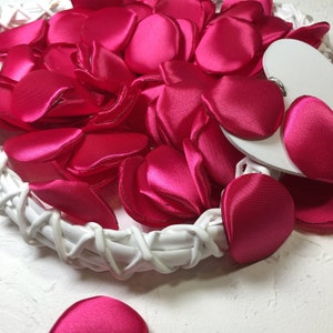 May include: Close-up of a white woven basket filled with bright pink satin rose petals. A white heart-shaped decoration is partially visible. The petals are scattered, creating a romantic and decorative display.