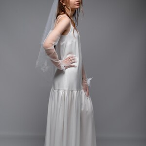 May include: A white satin wedding dress with a flowing skirt and spaghetti straps. The dress is paired with sheer gloves and a veil. The model is wearing white sandals. The dress has a simple, elegant design.