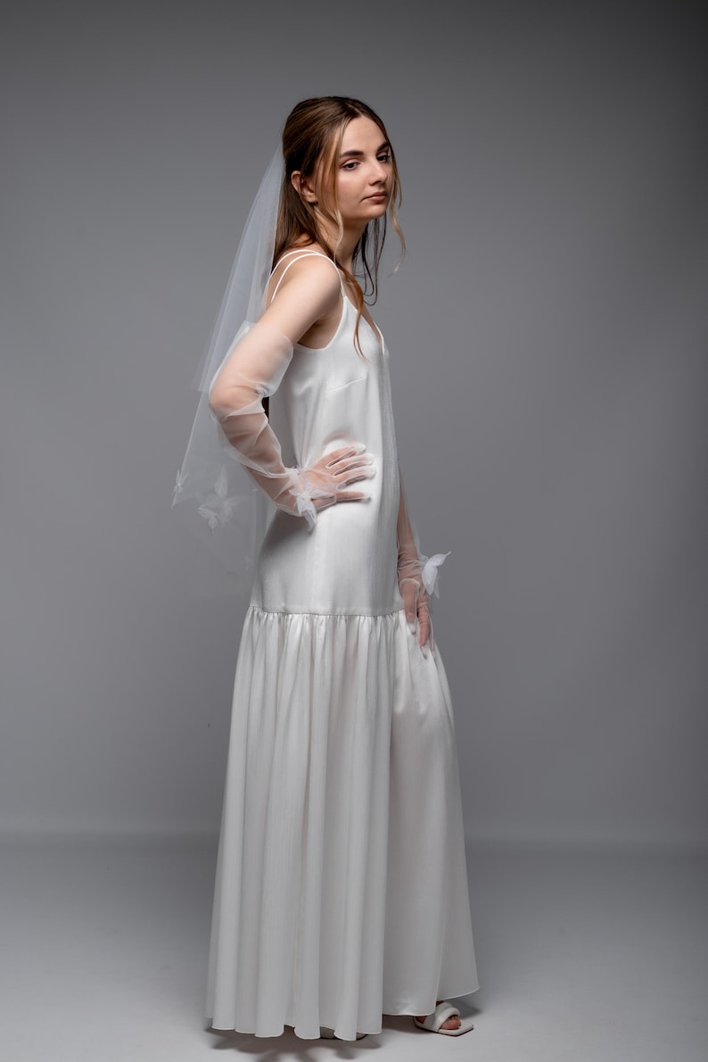 May include: A white satin wedding dress with a flowing skirt and spaghetti straps. The dress is paired with sheer gloves and a veil. The model is wearing white sandals. The dress has a simple, elegant design.