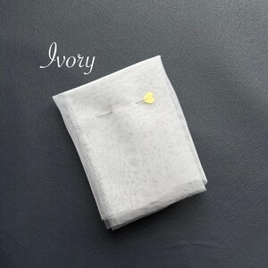 May include: Ivory-colored fabric, folded and secured with a yellow heart-shaped pin. The fabric is a light, sheer material. The word "Ivory" is written in cursive in the upper left corner. The fabric is on a dark gray surface.