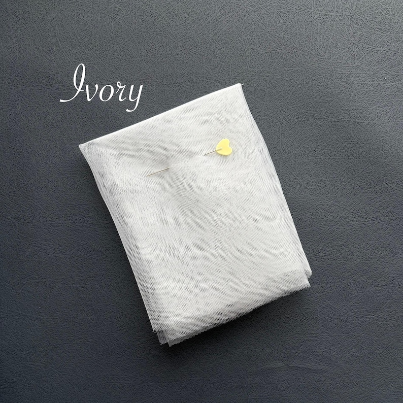 May include: Ivory-colored fabric, folded and secured with a yellow heart-shaped pin. The fabric is a light, sheer material. The word "Ivory" is written in cursive in the upper left corner. The fabric is on a dark gray surface.