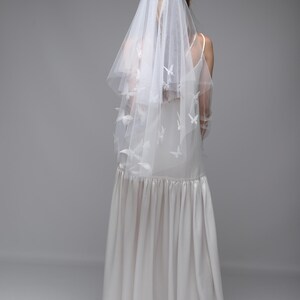 May include: A white wedding veil with butterfly accents. The veil is layered and drapes over a white dress. The dress has a fitted bodice and a flowing skirt. The background is a solid gray.
