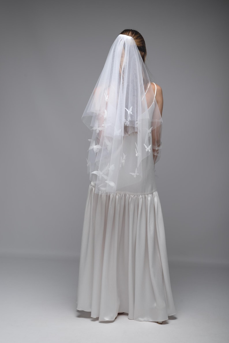 May include: A white wedding veil with butterfly accents. The veil is layered and drapes over a white dress. The dress has a fitted bodice and a flowing skirt. The background is a solid gray.