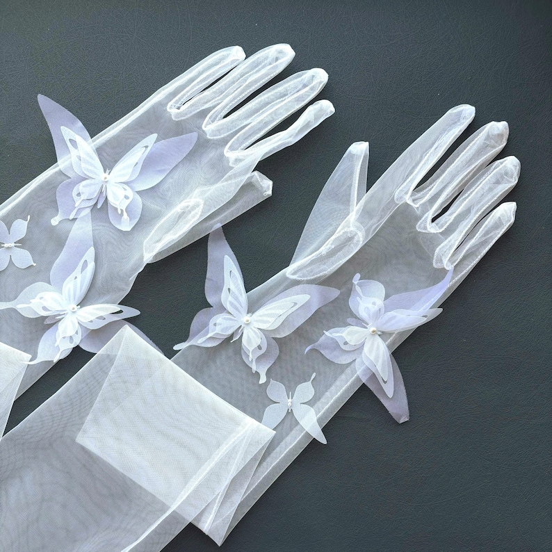 May include: A pair of sheer white gloves adorned with delicate white butterfly appliqu&eacute;s. The butterflies have detailed wings and are placed across the gloves. The gloves are made of a lightweight, see-through fabric.