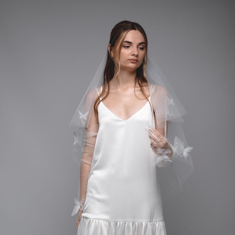 May include: A white bridal veil with butterfly accents. The veil is layered and appears to be made of a sheer fabric. The butterflies are white and add a delicate touch to the veil. The model is wearing a white dress.