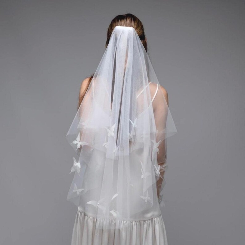 May include: A white bridal veil with butterfly accents. The veil is made of a sheer fabric and features multiple butterfly embellishments. The bride is wearing a white dress. The background is a solid gray.