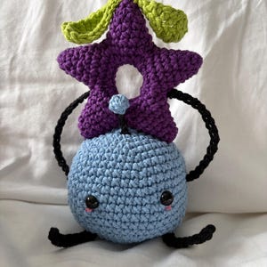 May include: A handmade crochet plush toy featuring a blue spherical body with a smiling face and black limbs. A purple star-shaped headpiece with green leaves sits atop the body. The toy is crafted with a tight stitch and detailed features.