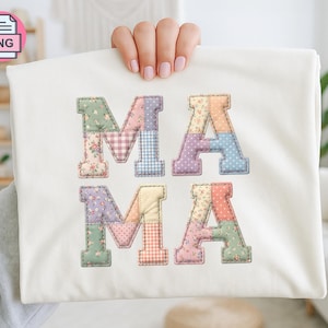 Può includere: Una maglietta bianca con la scritta "MAMA" in lettere patchwork. Le lettere sono realizzate con vari tessuti floreali e fantasia in colori pastello, tra cui rosa, verde, blu e giallo. La maglietta è tenuta da una persona.