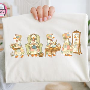 Puede incluir: Una camiseta color crema con una ilustración caprichosa de un ganso con un vestido de retazos, participando en varias actividades. El ganso se muestra con una cesta de flores, sentado en una silla, horneando y mirándose en un espejo. El diseño tiene una estética de libro de cuentos vintage.