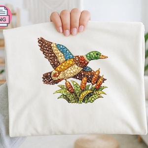 Pode incluir: Uma camiseta branca com uma ilustração detalhada de um pato em voo. O pato tem detalhes em marrom, azul, amarelo e verde, com um design detalhado. O pato está acima de um grupo de juncos verdes e amarelos.