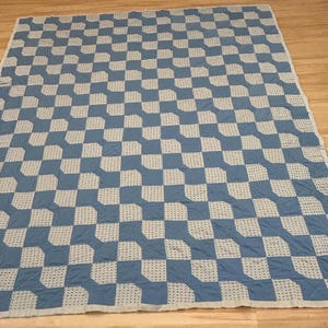 May include: A rectangular quilt featuring a repeating geometric pattern of solid blue squares and white crocheted squares. The design creates a checkerboard effect across the entire surface, finished with a simple white border along all four edges.