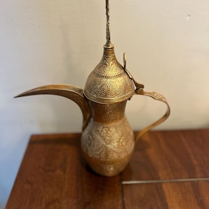 Puede incluir: Una cafetera de latón alta con un pico largo y curvado, una tapa con bisagras con un remate decorativo y un asa arqueada. La superficie exterior está cubierta con intrincados patrones florales y geométricos grabados en todo el cuerpo.