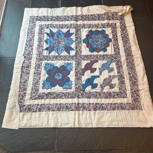 Può includere: Una trapunta fatta a mano con quattro diversi motivi quadrati. Ogni quadrato presenta un design floreale o geometrico unico nelle tonalità del blu e del viola, su uno sfondo color crema. La trapunta è bordata con una stampa floreale.
