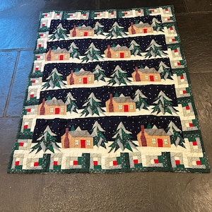 May include: A rectangular quilted blanket featuring a repeating pattern of tan cabins with red doors and chimneys, surrounded by green pine trees against a dark navy blue background with white speckles. The border consists of small green and red squares.