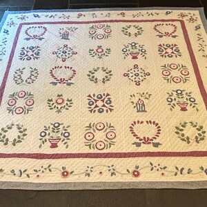 May include: Cream-colored quilt with a repeating pattern of floral wreaths, bouquets, and geometric designs in red, green, and blue. The quilt has a red border with floral accents and a blue edge.