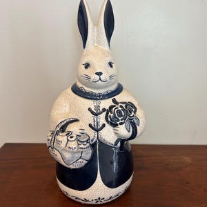 May include: A ceramic rabbit figurine with a cream-colored body and navy blue accents. The rabbit holds a bouquet of roses and a bag with text. The figurine has long ears and a decorative collar. The rabbit is approximately 12 inches tall.