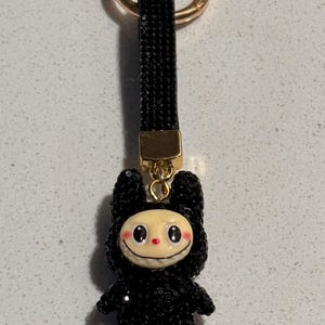 Pode incluir: Um porta-chaves preto coberto de strass com uma personagem de desenho animado num traje com capuz. A personagem tem um rosto sorridente com bochechas rosadas e um sorriso com dentes brancos. O porta-chaves tem um fecho dourado e uma correia preta.