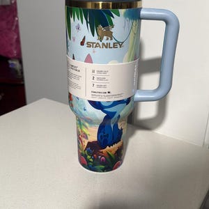 May include: A light blue Stanley tumbler with a handle and a tropical jungle print featuring a blue character. The cup has a gold rim, a clear lid with a light gray closure, and a white paper band displaying 11 hours cold, 2 days iced, and 7 hours hot.