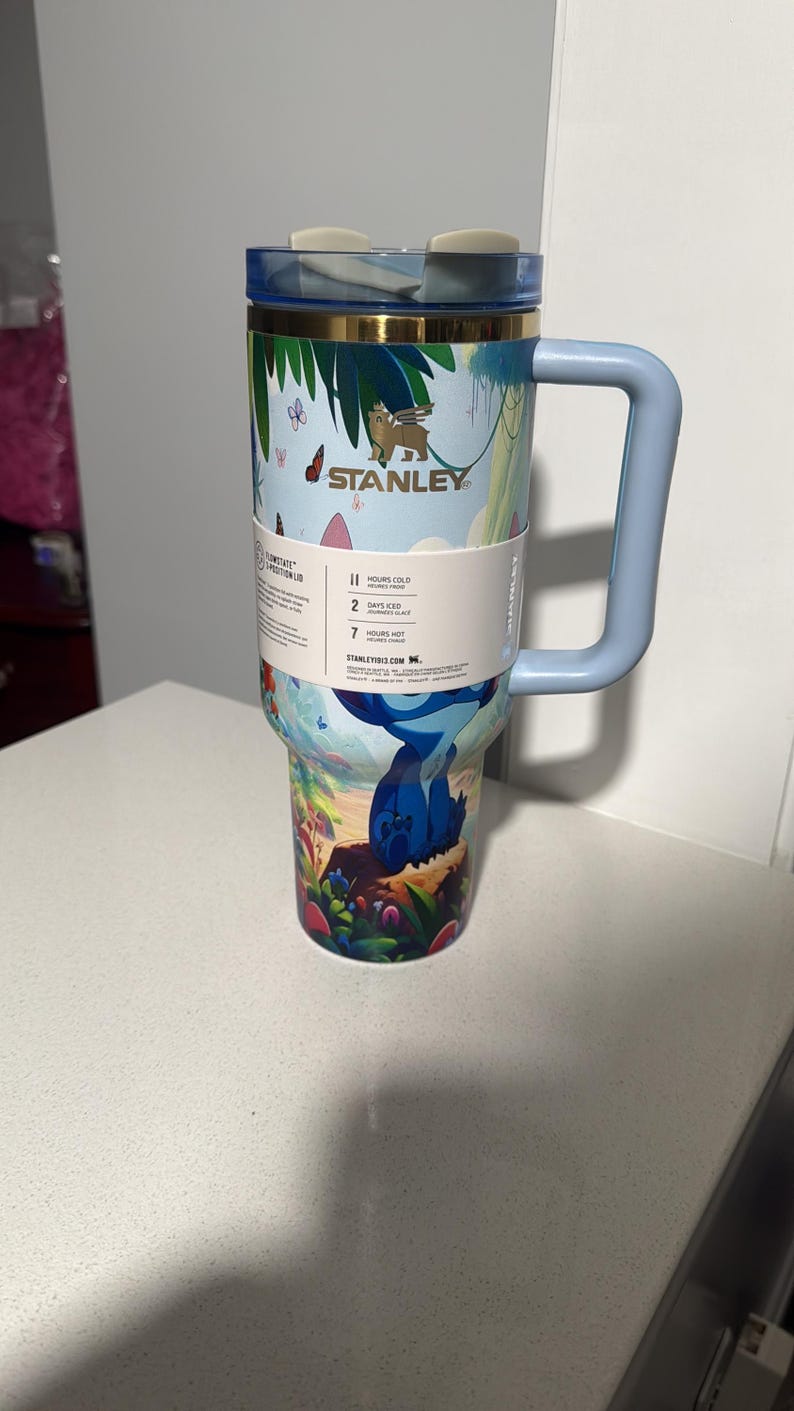 May include: A light blue Stanley tumbler with a handle and a tropical jungle print featuring a blue character. The cup has a gold rim, a clear lid with a light gray closure, and a white paper band displaying 11 hours cold, 2 days iced, and 7 hours hot.