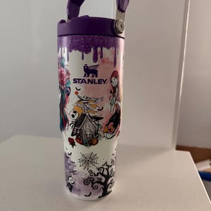 May include: A white Stanley tumbler featuring a purple lid and handle, decorated with a colorful graphic print of characters and motifs including a dog house labeled Zero, a spider web, and a tree, with purple paint drip accents at the top.