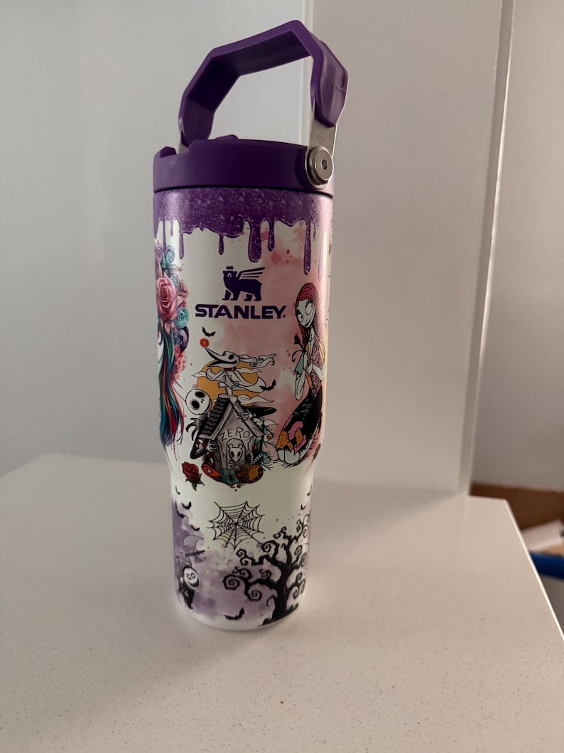 May include: A white Stanley tumbler featuring a purple lid and handle, decorated with a colorful graphic print of characters and motifs including a dog house labeled Zero, a spider web, and a tree, with purple paint drip accents at the top.