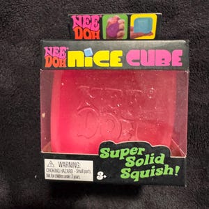 May include: A red Nee Doh Nice Cube sensory toy packaged in a black box with a clear plastic window. The packaging features the text Nee Doh Nice Cube and Super Solid Squish, along with a warning label regarding small parts for children under 3.