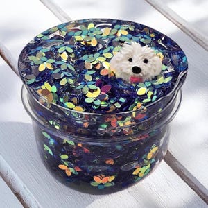 May include: A clear plastic container filled with dark blue glitter and flower-shaped sequins. A white, fluffy dog figurine with a pink tongue is on top of the glitter. The container is sitting on a white surface.