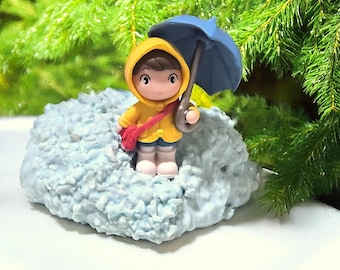 May include: A small figurine of a character wearing a yellow hooded raincoat and white boots, holding a blue umbrella and carrying a red crossbody bag. The figure stands on a textured, light blue base that resembles a cloud or pile of snow.